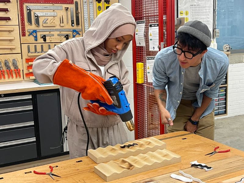 A girl wearing a hijab and grey hoodie standing beside a man wearing a grey hat, glasses, and a blue shirt. The girl is holding a large industrial tool and burning a piece of wood while the man observes her work. 